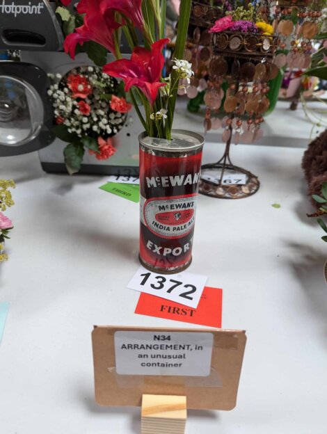 A can labeled "McEwan's Export" is used as a vase for a floral arrangement. The display is marked with the number 1372 and a "first" place label. A sign reads, "N34 ARRANGEMENT, in an unusual container.