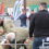 A group of people examine and handle sheep in a pen at an outdoor event. Flags and booths are visible in the background.