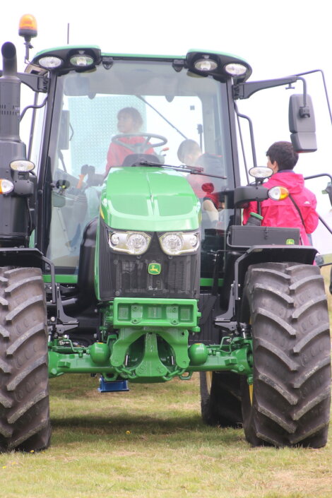 A green tractor parked on grass with children seated inside the cab and an adult standing beside it.