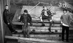 Black and white photo of four men outdoors, casually standing and sitting on stacks of wooden pallets and construction debris, with an old metal bed frame in the background.