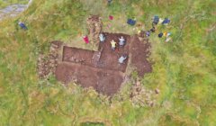 Aerial view of an archaeological excavation site with several people working in a cleared earth area surrounded by grassy terrain. Various tools and bags are scattered around the site.