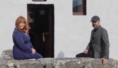 Two people sit on a stone wall in front of a white building with a doorway and window. The person on the left wears a blue dress, and the person on the right wears a gray jacket and a cap.