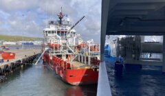 A red and white supply vessel is docked at a port next to a concrete pier, with various equipment and personnel visible on board. The sky is partly cloudy.