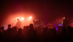 A band performs on stage with guitars and drums under orange and blue lighting as a crowd watches in the foreground.