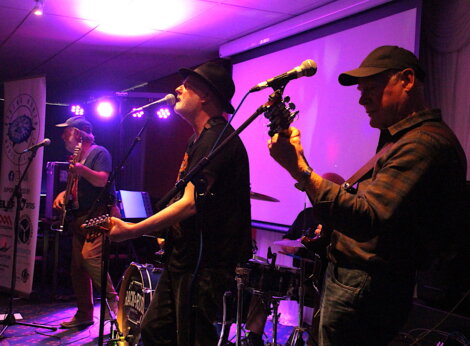 A band of four musicians performs live on stage under purple lighting. Two guitarists, a bassist, and a drummer are visible. A large screen and various music equipment are in the background.