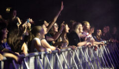 A crowd of people stand behind a barrier at a concert. Some are raising their hands and cheering, while others are looking towards the stage. The area is dimly lit with concert lighting.