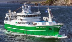 A green and white fishing vessel named "Antarctic" travels through a body of water with rocky cliffs and hills in the background. The vessel is adorned with international maritime signal flags.