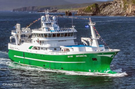 A green and white fishing vessel named "Antarctic" travels through a body of water with rocky cliffs and hills in the background. The vessel is adorned with international maritime signal flags.