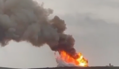 A large plume of smoke and flames is seen rising from a hill, indicating a significant fire or explosion with cloudy skies in the background.