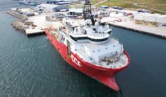 A large red and white research vessel is docked at an industrial harbor, with several buildings, shipping containers, and equipment in the background.