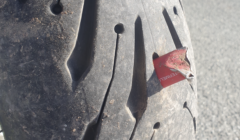 A close-up image of a tire with a sharp metal piece embedded in the tread, causing visible damage. The tire has a textured surface, and the background shows a paved road.