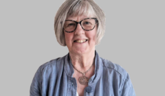 A woman with short gray hair and glasses is smiling. She is wearing a blue top and a necklace with a circular pendant. The background is gray.