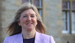 A woman with long blond hair smiles, wearing a light purple blazer, standing in front of a building with stone walls and windows.