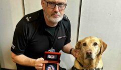 A man in uniform holds a medal while kneeling beside a Labrador retriever wearing a harness. They both face the camera inside an indoor setting.