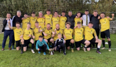 A soccer team in yellow jerseys poses for a group photo on a field with a trophy, celebrating a victory. Two children in front and coaches in suits stand behind the players.