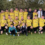 A soccer team in yellow jerseys poses for a group photo on a field with a trophy, celebrating a victory. Two children in front and coaches in suits stand behind the players.