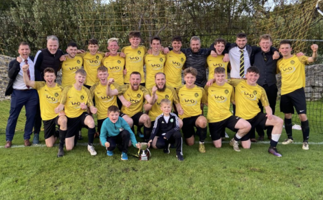 A soccer team in yellow jerseys poses for a group photo on a field with a trophy, celebrating a victory. Two children in front and coaches in suits stand behind the players.