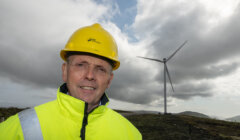 A man in a yellow hard hat and high-visibility jacket stands in a hilly area with a wind turbine in the background under a cloudy sky.