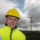 A man in a yellow hard hat and high-visibility jacket stands in a hilly area with a wind turbine in the background under a cloudy sky.