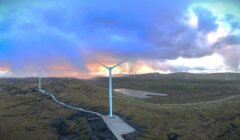 A wind farm with multiple wind turbines stands on a vast, hilly landscape under a partly cloudy sky at sunset.