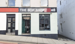 Street view of "The Bop Shop," a storefront with large windows and a black sign with white text above the entrance door. The surrounding buildings are white and grey.