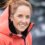 A woman with light brown hair tied up, wearing an orange hooded jacket, smiles at the camera with a blurred outdoor background.