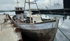 A weathered, wooden fishing boat tied to a concrete pier with a larger vessel in the background on a cloudy day.