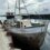 A weathered, wooden fishing boat tied to a concrete pier with a larger vessel in the background on a cloudy day.