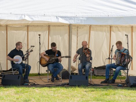 Four musicians perform under a white tent, playing a banjo, guitar, violin, and accordion. They are seated in a row with microphones and amplifiers set up before them.