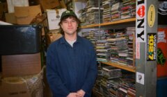 Person standing in a cluttered room, surrounded by stacks of CDs and boxes. The individual is wearing a blue jacket. Shelving units are seen in the background with more CDs and miscellaneous items.