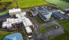 Aerial view of a school complex with multiple connected buildings, parking areas, surrounding fields, and sports facilities including a football pitch. Paths and roads run through the premises.