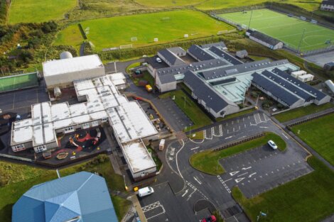 Aerial view of a school complex with multiple connected buildings, parking areas, surrounding fields, and sports facilities including a football pitch. Paths and roads run through the premises.