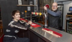 Two people are in a bar. The woman is seated at the bar counter, and the man is standing behind it, with beer taps visible in front of them.