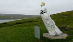 A statue of a woman in a white dress, leaning into the wind, stands on a grassy hill with a sign nearby, overlooking water and distant hills.