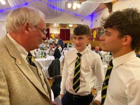 Three men, two younger and one older, are engaged in conversation at an indoor event with tables set for dining in the background. They are all wearing formal attire, including white shirts and striped ties.