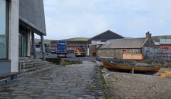 Waterside scene depicting a stone walkway, a docked wooden boat, and an industrial area with blue shipping containers and a forklift in the background.