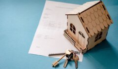 A small model house, document, and a set of keys are arranged on a blue surface.