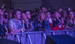 A group of people stand at a barricade, watching an event. Some are holding drinks, and others are taking photos. The area is dimly lit with purple lighting.