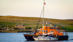 An orange RNLI lifeboat assists a smaller sailboat in a calm body of water, with a grassy hillside and buildings in the background.