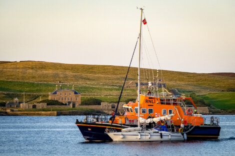 An orange RNLI lifeboat assists a smaller sailboat in a calm body of water, with a grassy hillside and buildings in the background.