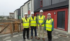 Four people wearing yellow safety vests and white helmets stand in front of newly constructed homes.