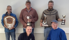 Five men pose with trophies and a shield. One man holds a large cup, another holds a smaller cup and shield, another holds a trophy with multiple small shields. All are smiling and facing the camera.