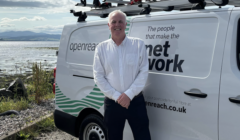 A man stands in front of a white van with "Openreach" branding. The van is parked on a gravel area near a large body of water.