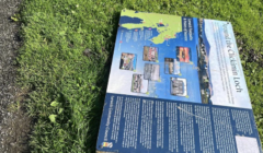 An informational sign detailing local attractions and history lies on the grass beside a paved path.