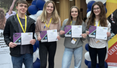 Four young individuals stand side by side holding certificates, wearing lanyards, and smiling in front of a backdrop with balloons.