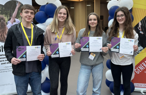 Four young individuals stand side by side holding certificates, wearing lanyards, and smiling in front of a backdrop with balloons.