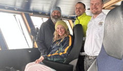 Four people onboard a boat's bridge, smiling at the camera. One person is seated while the others stand behind. The background shows the interior of the bridge with large windows.