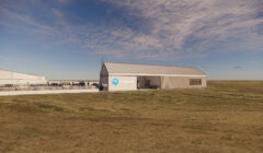 A single-story building with a "Statkraft" logo on the facade is situated in an open field under a partly cloudy sky. Several parked cars are visible in the background.