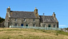 A stone house with two chimneys and a slate roof stands on a grassy hill, surrounded by a light blue picket fence under a clear blue sky.