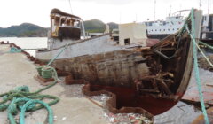 An old, damaged wooden boat is docked at a pier. The hull appears heavily deteriorated, with visible rot and missing planks. Green ropes are strewn around the dock.